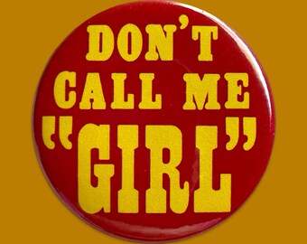 Puede incluir: Un botón redondo rojo con el texto "DON'T CALL ME "GIRL"" en letras amarillas y en negrita. El botón tiene un acabado brillante y está sobre un fondo amarillo mostaza. El texto está centrado y es fácil de leer.