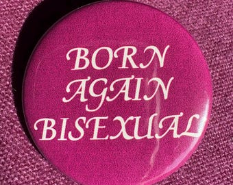 May include: A round, magenta button with the text "BORN AGAIN BISEXUAL" in white, stylized lettering. The button is set against a textured, magenta background, creating a bold contrast.