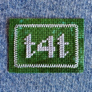 May include: A rectangular green fabric patch featuring the embroidered light gray text t4t in the center. The patch is bordered by a white stitched rectangular frame and is set against a textured blue denim fabric background.