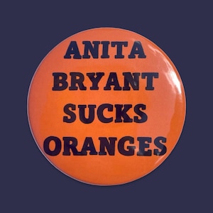May include: A round, orange button with the text "ANITA BRYANT SUCKS ORANGES" in bold, black capital letters. The button has a glossy finish and is set against a dark blue background.