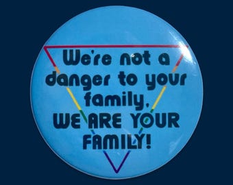 Puede incluir: Un botón redondo azul claro con el texto "We're not a danger to your family, WE ARE YOUR FAMILY!" en azul oscuro. Un triángulo arcoíris y un triángulo rojo enmarcan el texto.
