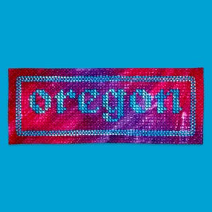 May include: A rectangular cross-stitch embroidery piece featuring the word Oregon in light blue letters. The background is a variegated blend of red, purple, and pink threads, enclosed within a light blue stitched rectangular border.
