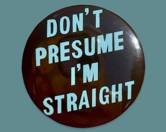 Puede incluir: Un botón redondo negro con el texto "DON'T PRESUME I'M STRAIGHT" en azul claro. El botón tiene un acabado brillante y está sobre un fondo turquesa.