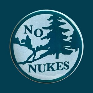 May include: Round button with a teal border and a teal pine tree silhouette. The words "NO NUKES" are printed in teal, with "NO" above the tree and "NUKES" below. The background is a light, textured silver.