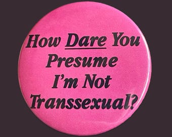 May include: A circular, vibrant pink button with black lettering. The text on the button says: "How Dare You Presume I'm Not Transsexual?" The button has a shiny surface and is set against a dark backdrop.