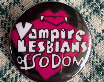 Op de afbeelding: Een ronde button met een donkere achtergrond en een magenta hartvorm in het midden. De tekst "Vampire LESBIANS of SODOM" is in het wit over het hart gedrukt, dat aan de bovenzijde in het midden is voorzien van twee kleine witte hoektanden.