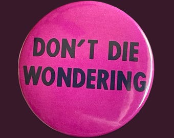 Puede incluir: Un botón redondo de color rosa intenso con la frase "DON'T DIE WONDERING" impresa en letras negras y en negrita. El botón tiene un acabado brillante y está sobre un fondo morado oscuro.