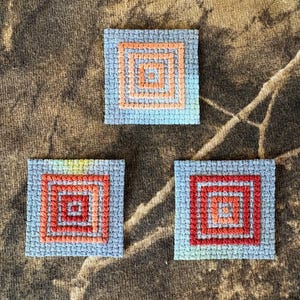 May include: Three square fabric patches featuring cross-stitch embroidery. Each patch has a light blue background with a series of concentric squares in shades of orange and red stitched in the center, creating a geometric pattern on each piece.