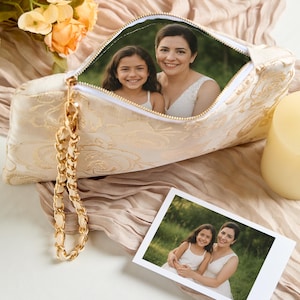 May include: A cream-colored clutch with a gold floral pattern and a gold chain wrist strap. The clutch is open, revealing a photo of a woman and a young girl. A second photo of the same pair is next to the clutch.
