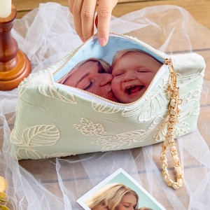 May include: A light green clutch bag featuring white floral embroidery and a gold chain wrist strap. The interior lining displays a printed photograph of an adult kissing a laughing child on the cheek, matching a separate photo print placed below.