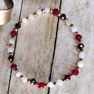 May include: A beaded bracelet featuring alternating white, burgundy, and dark red faceted beads. The beads are separated by small gold-colored beads. The bracelet is displayed on a light brown wooden surface.