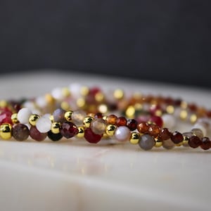 May include: A beaded bracelet featuring small, faceted, multi-colored gemstone beads in shades of red, brown, white, and grey, interspersed with small, polished, round gold-tone metal spacer beads arranged in a continuous circular pattern.
