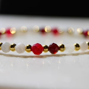 May include: A beaded bracelet featuring a repeating pattern of faceted red beads, smooth white translucent beads, and small polished gold-tone metallic spherical beads arranged in a circular band design.