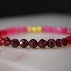 May include: A beaded bracelet featuring a gradient of faceted dark red garnet-colored beads transitioning into bright pink beads, accented with three small, smooth, round gold-toned metallic beads positioned at the center of the strand.