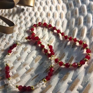 May include: A close-up of two beaded necklaces. One necklace features faceted red beads and gold-colored accents. The other necklace has clear, faceted beads and gold-colored accents. Both necklaces are on a white surface.