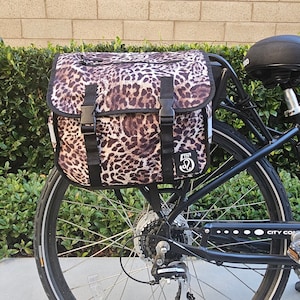 Könnte beinhalten: Ein Fahrrad mit einer Gepäckträgertasche mit Leopardenmuster, die am Gepäckträger befestigt ist. Die Tasche ist braun und schwarz mit schwarzen Riemen und Schnallen. Das Fahrrad ist schwarz mit einem schwarzen Sattel und einem silbernen Rad.