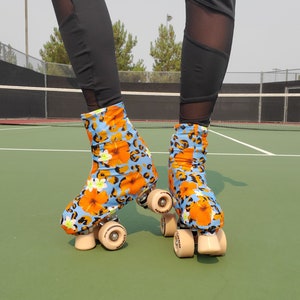 Op de afbeelding: Roller skates met een blauw bloemenpatroon en oranje bloemen. De skates hebben beige wielen en staan op een groene ondergrond. De persoon draagt ​​zwarte leggings.