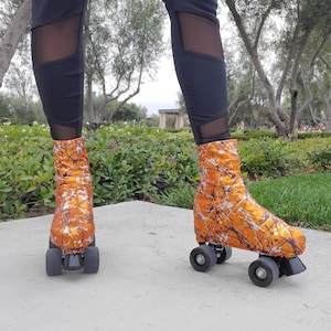 May include: Orange roller skate covers with a white and black splatter design. The covers are on black roller skates with grey wheels. The person is wearing black leggings with mesh panels.