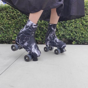 Op de afbeelding: Paar rolschaatsen met zwart-witte laarzen met marmerpatroon. De rolschaatsen hebben grijze wielen en zwarte stoppers. De persoon draagt een lange zwarte rok. De achtergrond is een groene haag en een grijze stoep.