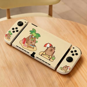 Puede incluir: Una consola Nintendo Switch de color crema con un diseño de temática playera. La consola presenta ilustraciones de capibaras de dibujos animados relajándose en la playa, usando gafas de sol y disfrutando de cocos. Los mandos tienen botones negros.