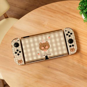May include: A Nintendo Switch console with a brown and white checkered design. The console features a cute bear illustration with a party hat and bow tie, along with the text "Cute Bear". The Joy-Cons also have bear and strawberry details.