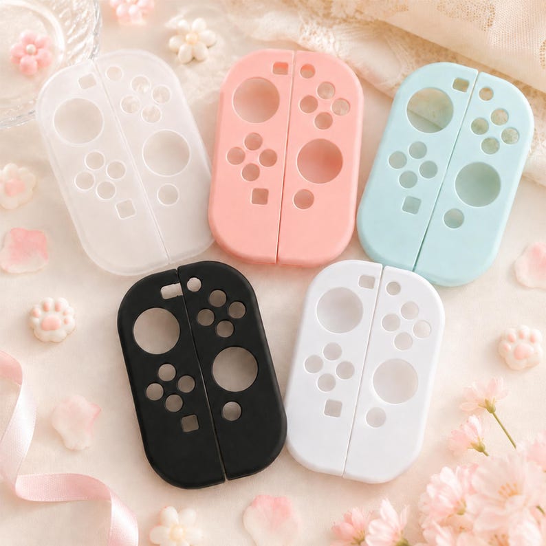May include: A collection of five colored controller cases. The cases are in white, pink, light blue, black, and white. Each case is designed to fit a gaming controller, with button and joystick cutouts. The cases are arranged on a light-colored surface.