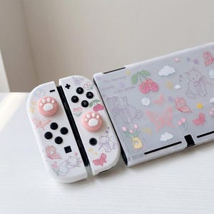 Puede incluir: Mandos Joy-Con blancos de Nintendo Switch y una funda transparente decorada con mariposas rosas, gatos y cerezas. Los mandos tienen agarres para el pulgar rosas con forma de pata. La funda tiene el texto "The charming scenery in the garden."