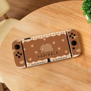 Puede incluir: Una consola Nintendo Switch marrón y blanca con un diseño personalizado. El diseño presenta un capibara durmiendo, corazones y la palabra "KAPIBALA". La consola está sobre una mesa de madera.