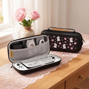 May include: Black and white gaming console case with a handle, open to reveal the console and accessories. A second black case with a bunny and strawberry pattern is next to it. Pink roses in a vase are in the background.