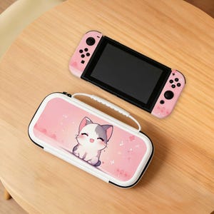 May include: A pink Nintendo Switch console and matching carrying case. The Switch has pink controllers with floral accents. The case features a cartoon cat illustration on a pink background, with a white handle and zipper.