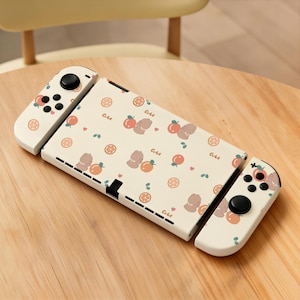 May include: A portable gaming console featuring a cream-colored protective skin with a repeating pattern of small brown bears, orange fruit slices, and the word "Cute" in a cursive font, complete with matching cream-colored side controllers.