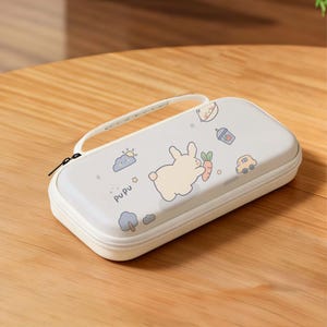 May include: A light gray, rectangular case with a white zipper and handle. The case features cartoon illustrations of a bunny, carrot, car, cloud, and the word "pupu". The case is on a wooden surface.