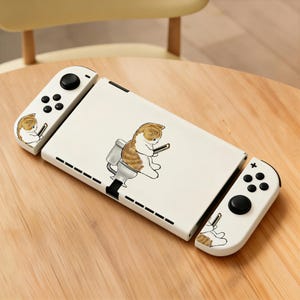 May include: White Nintendo Switch console with a custom design featuring a cartoon cat on a toilet, holding a phone. The controllers also have a matching cat design. The console is on a wooden surface.