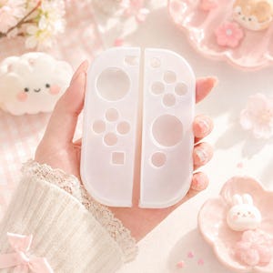 May include: Two white Nintendo Switch Joy-Con controllers. The controllers are held in a hand, with a soft, blurred background of pink flowers and decorative elements. The controllers have a translucent appearance, with visible button outlines.