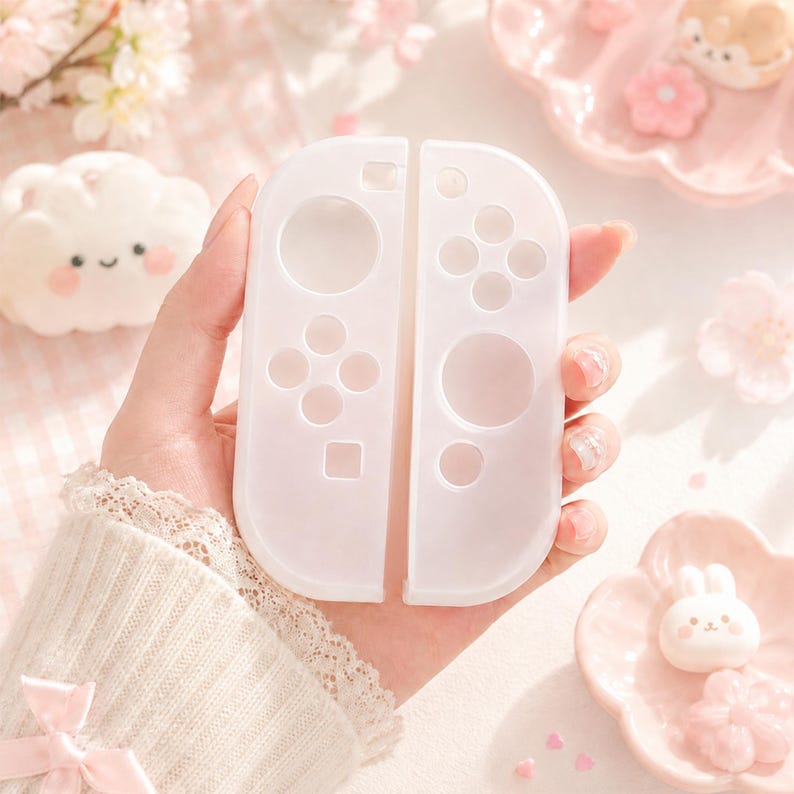 May include: Two white Nintendo Switch Joy-Con controllers. The controllers are held in a hand, with a soft, blurred background of pink flowers and decorative elements. The controllers have a translucent appearance, with visible button outlines.