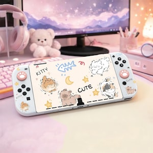 Puede incluir: Una consola Nintendo Switch blanca con un diseño personalizado con gatos de dibujos animados, estrellas y la palabra "CUTE". Los mandos tienen huellas de patas rosas y la palabra "KITTY". La consola está sobre un escritorio rosa.