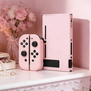 May include: A pink games console and controller set. The console is rectangular, with a black panel at the bottom. The controller is split into two parts, each with black buttons and joysticks. A bouquet of pink flowers sits in the background.