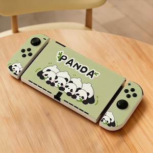 May include: A light green gaming console case with the word "PANDA" and a graphic of panda bears. The case has two detachable controllers, each with black buttons and a panda graphic. The console is on a wooden surface.