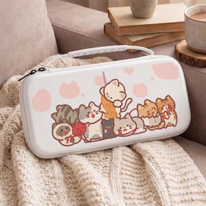 以下が含まれることがあります： 白いキャリングケース、漫画の猫のデザイン。ケースにはハンドルとジッパー開閉部が付いています。デザインには、さまざまな猫、糸、ピンクの足跡が含まれています。ケースはクリーム色のニットブランケットの上に置かれています。