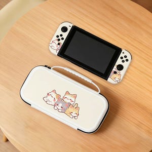Può includere: Una console Nintendo Switch bianca e una custodia abbinata, entrambe decorate con simpatiche illustrazioni di gatti dei cartoni animati. La console ha uno schermo nero e controller bianchi. La custodia ha una maniglia e una cerniera.