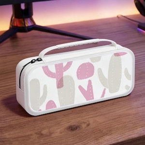 Pode incluir: Estojo retangular branco com alça e zíper preto. O estojo apresenta um padrão de cactos rosa e cinza em um fundo branco. O estojo está em uma superfície de madeira.