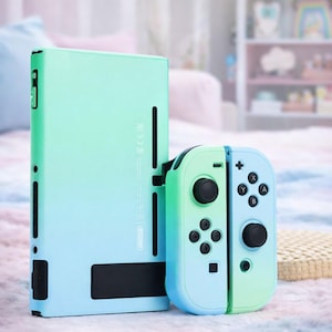 Pode incluir: Uma consola de jogos com gradiente verde-menta e azul com comandos a condizer. A consola e os comandos são mostrados juntos, com os comandos desligados. Os comandos têm botões pretos e almofadas direcionais.