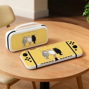 May include: A yellow gaming console and a matching rectangular carrying case, both featuring a graphic illustration of three cats—one gray, one white, and one black—enclosed within a delicate floral frame on a solid yellow background.