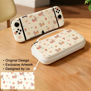 Puede incluir: Una consola de juegos portátil y un estuche de transporte rectangular a juego, ambos con una superficie de color crema con un patrón repetitivo de pequeños osos marrones, rodajas de naranja, hojas verdes y la palabra "Cute" en cursiva.