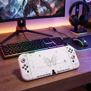 以下が含まれることがあります： 白いゲーム機。蝶の羽のデザイン、足跡のボタン、星のアクセントが特徴です。レインボーライトのキーボード、マウス、ヘッドセットも木製の机にあり、ゲーム環境を示唆しています。