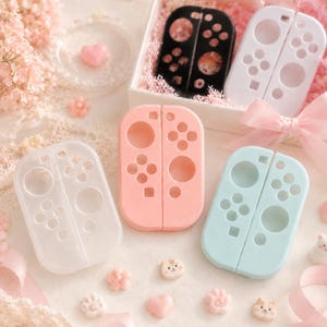 May include: A collection of pastel-colored gaming controller accessories. The image features pink, blue, white, and clear controller shells, along with decorative elements like heart-shaped and floral embellishments. Some controllers are in a gift box.