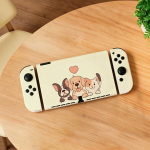 Op de afbeelding: Een crèmekleurige spelcomputer met een afbeelding van een Franse bulldog, een golden retriever en een kat met een roze hartje erboven. De console heeft aan weerszijden witte afneembare controllers met zwarte knoppen en joysticks.