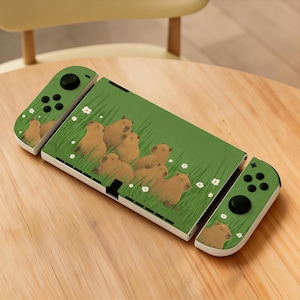 Puede incluir: Una consola Nintendo Switch verde y blanca con un diseño de carpincho. La consola presenta un fondo verde con ilustraciones de carpinchos, hierba y flores blancas. Los mandos son verdes con botones negros.