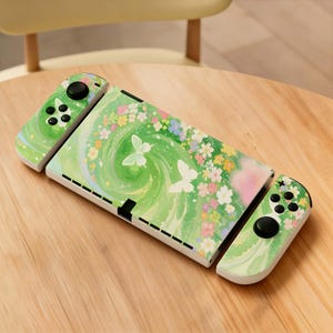 May include: A green and white Nintendo Switch console with matching Joy-Cons. The console features a floral and butterfly design with a green swirl background. The Joy-Cons have black control buttons and a green and white pattern.