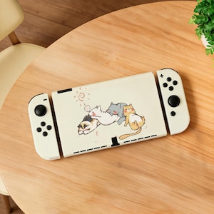 May include: A white gaming console dock featuring a graphic illustration of three cartoon cats cuddling together. The dock is flanked by two white controllers with black buttons and joysticks, displaying a clean and simple aesthetic design.
